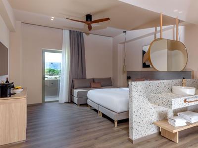 Chambre d'hôtel moderne avec lit double, canapé, grande douche et balcon donnant sur la verdure.