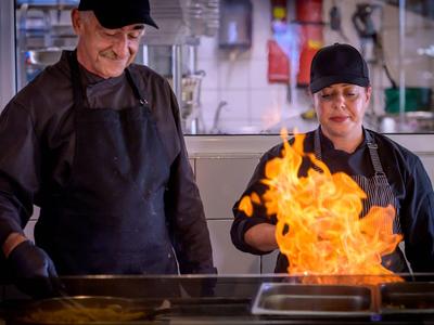 Zwei Köche in schwarzen Uniformen arbeiten an einer Pfanne mit großem Feuer im Restaurant.