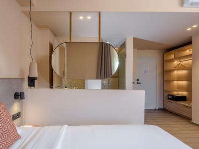 Modernes Hotelzimmer mit weißem Bett, rundem Spiegel und offenem Kleiderschrank mit Beleuchtung.