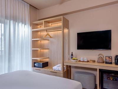 Hell eingerichtetes Hotelzimmer mit offenem Kleiderschrank, TV und Mini-Bar.