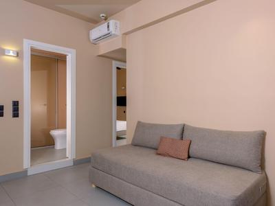 Modernes Hotelzimmer mit grauem Schlafsofa, kleinem Kissen und offener Badezimmertür.