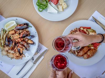 Stůl se dvěma talíři grilované ryby a salátu, ruce přiťukávající si sklenicemi červeného vína.