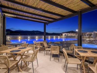 Überdachte Außenterrasse mit Holzstühlen und Tischen neben einem beleuchteten Pool in der Abenddämmerung.