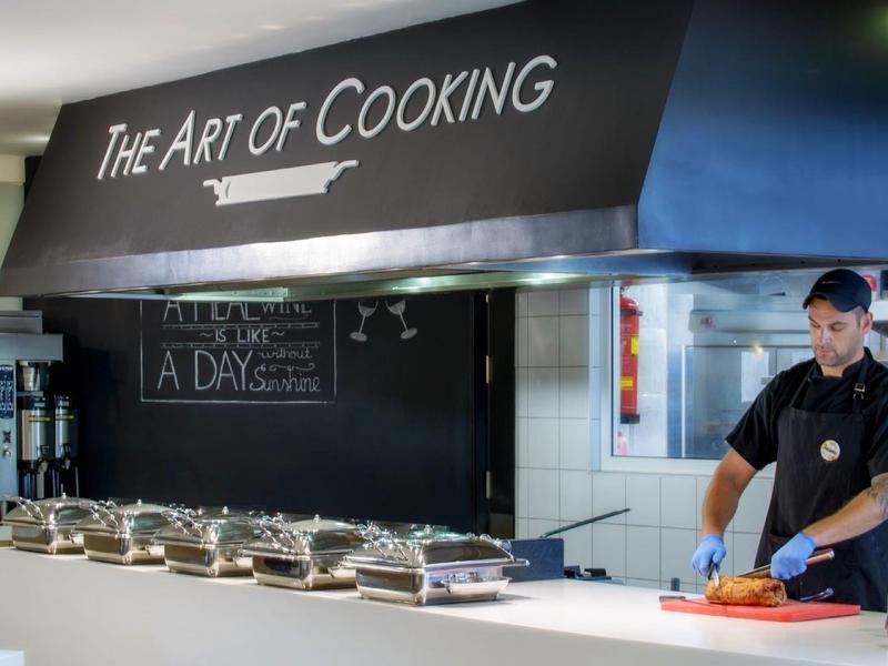 Kuchař připravuje jídlo v bufetové kuchyni pod nápisem ‚The Art of Cooking‘.