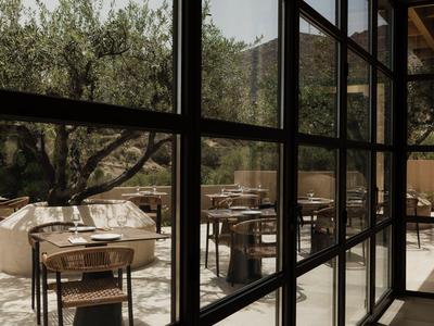 Nowoczesny zewnętrzny teren wypoczynkowy z krzesłami i stołami widoczny przez duże okna w czarnych ramach.