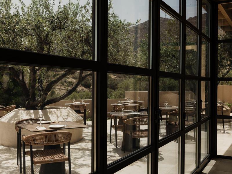 Buitenzitgedeelte met houten stoelen en ronde tafels gezien door grote glazen ramen.