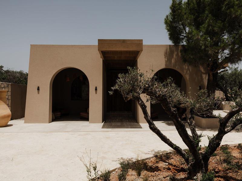 Modern gebouw in woestijnstijl met bogen, een boom en aardse kleuren onder een heldere lucht.