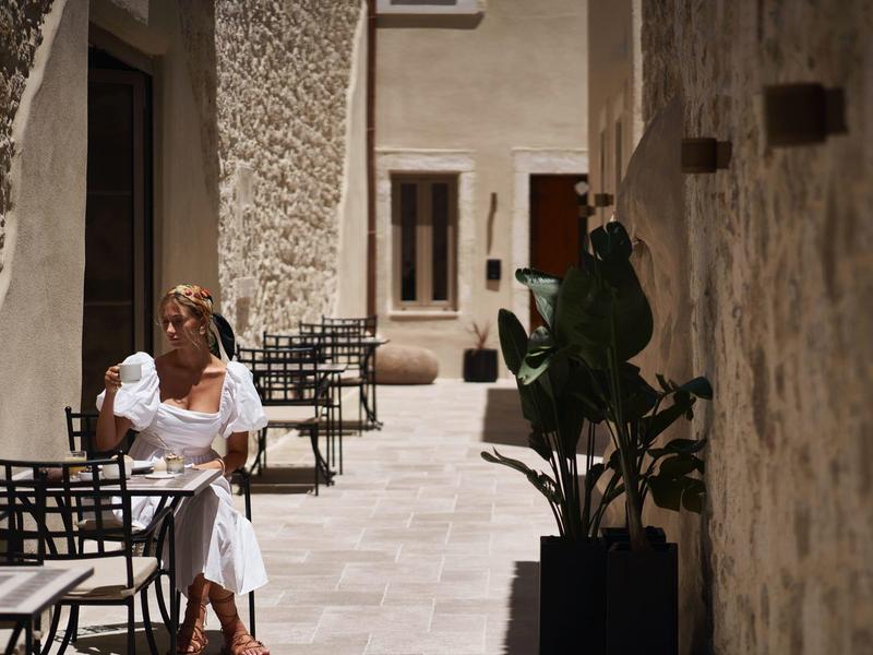 Femme en robe blanche assise dans une cour étroite baignée de soleil avec des murs en pierre.