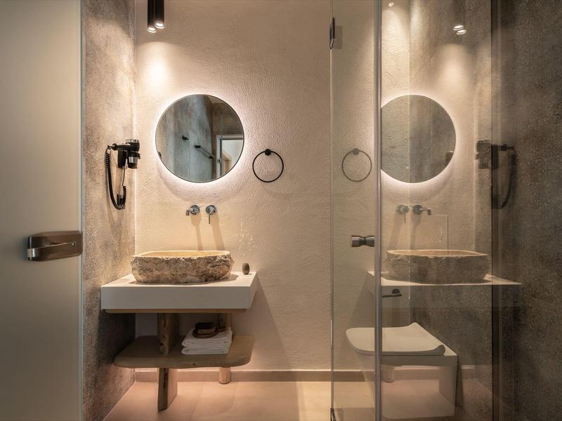 Salle de bain d'hôtel moderne avec miroirs ronds, lavabo en pierre et cabine de douche séparée.