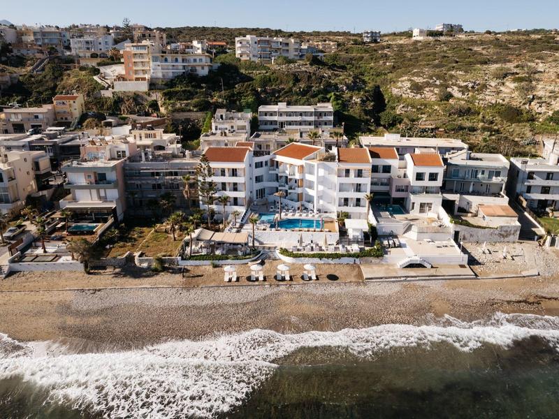 Hotel nadmorski z basenem i leżakami na plaży, otoczony apartamentami i wzgórzami.