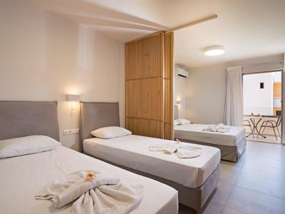 Jasny pokój hotelowy z dwoma łóżkami pojedynczymi, jednym podwójnym oraz balkonem z stołem i krzesłami.