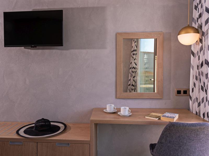 Nowoczesne biurko w pokoju hotelowym z lustrem, telewizorem, krzesłem, lampą, zestawem do herbaty i dekoracyjną zasłoną.