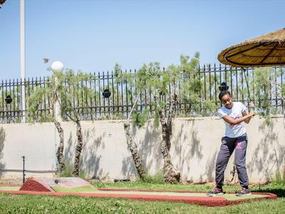 Uomo gioca a minigolf su un campo soleggiato con ombrelloni e recinzione sullo sfondo
