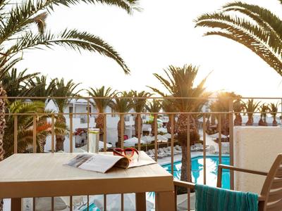 Balcone con tavolo, sedia e bevanda con vista sulla piscina e palme al tramonto.