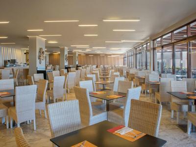 Světlá, moderní hotelová restaurace s mnoha stoly a židlemi, velkými okny a teplým dřevěným designem.