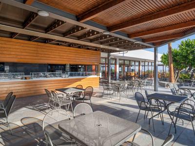 Venkovní terasa s stolky, židlemi a dřevěným obložením za slunečného počasí.