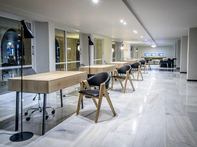 Moderní pracovní prostor v hotelu s dlouhými stolky, černými židlemi a světlou mramorovou podlahou.