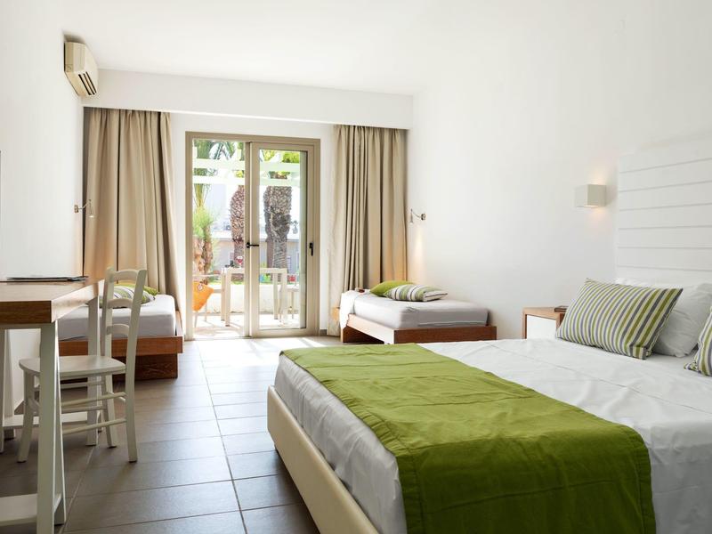 Světly hotelový pokoj se třemi postelemi, zeleným přehozem a přístupem na balkon.