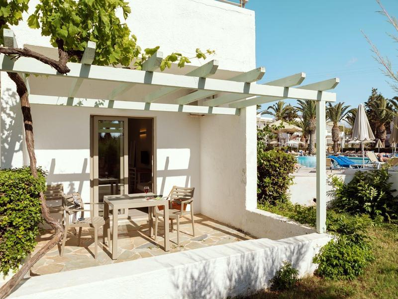 Slunná terasa s dřevěnými židlemi a stolem, obklopená zelení a výhledem na bazén.