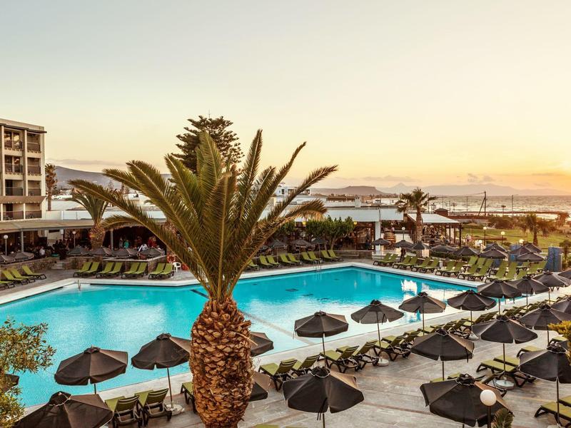 Hotelový bazén s palmami a lehátky při západu slunce s výhledem na moře v pozadí.