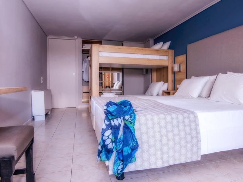 Hotelový pokoj s manželskou postelí, palandou, židlemi a modrým šátkem na posteli.