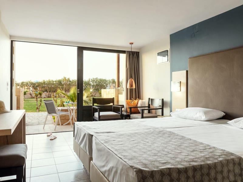 Světý hotelový pokoj s manželskou postelí, židlemi a velkým oknem s výhledem na zelenou krajinu.