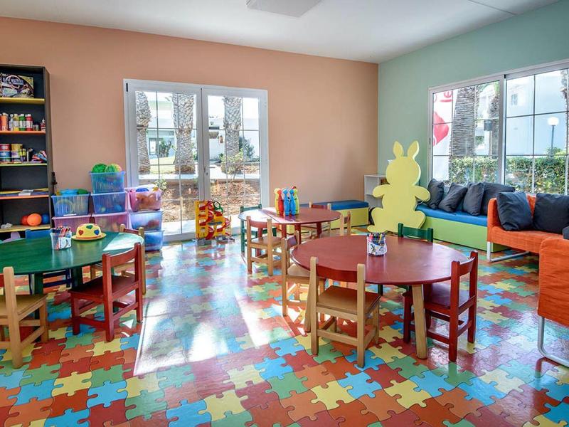 Barevná dětská herna s malými stolky, židlemi a hračkami na podlaze z puzzle koberců.