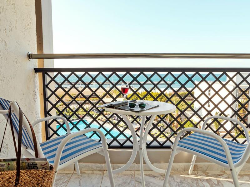 Balkon s malým bílým stolkem a dvěma židlemi s modro-bílými polštáři, výhled na moře.