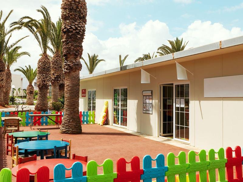 Venkovní prostor hotelu s barevným plotem, stoly a palmami za slunečného počasí