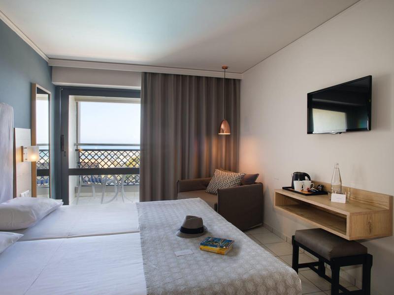 Moderně zařízený hotelový pokoj s manželskou postelí, posezením a balkonem s výhledem na moře.