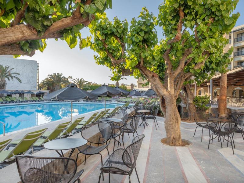 Venkovní hotelová oblast s bazénem, zelenými židlemi a stromy.