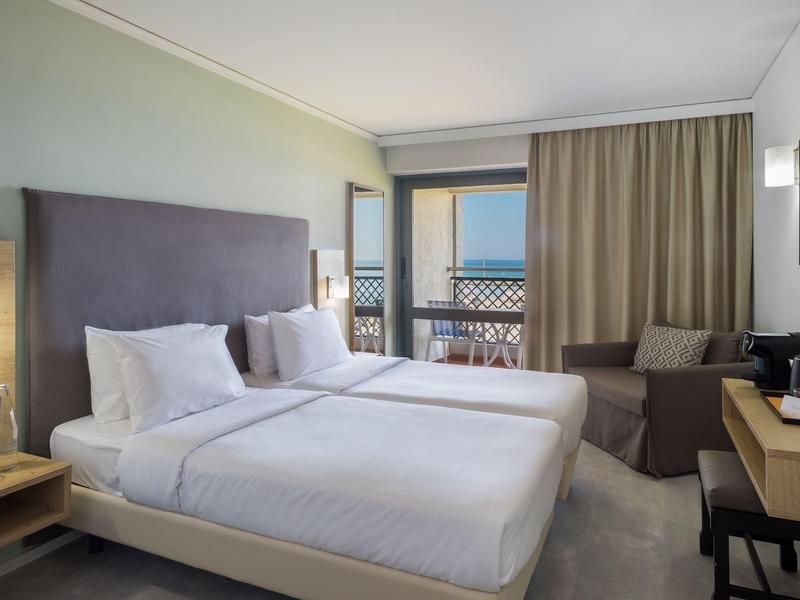 Moderní hotelový pokoj se dvěma lůžky, balkonem s výhledem na moře a útulným posezením.