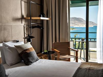 Przytulny pokój hotelowy z łóżkiem, drewnianym krzesłem i balkonem z widokiem na morze i góry.