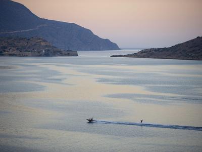 Łódź płynie po spokojnym morzu z odległymi wzgórzami o zachodzie słońca.