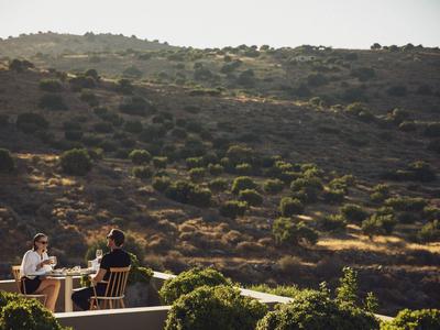 Dwie osoby jedzące na zewnątrz z widokiem na nasłonecznione wzgórze pokryte krzewami.