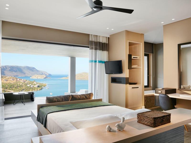 Nowoczesny pokój hotelowy z łóżkiem, telewizorem, komodą i dużym oknem z widokiem na ocean.