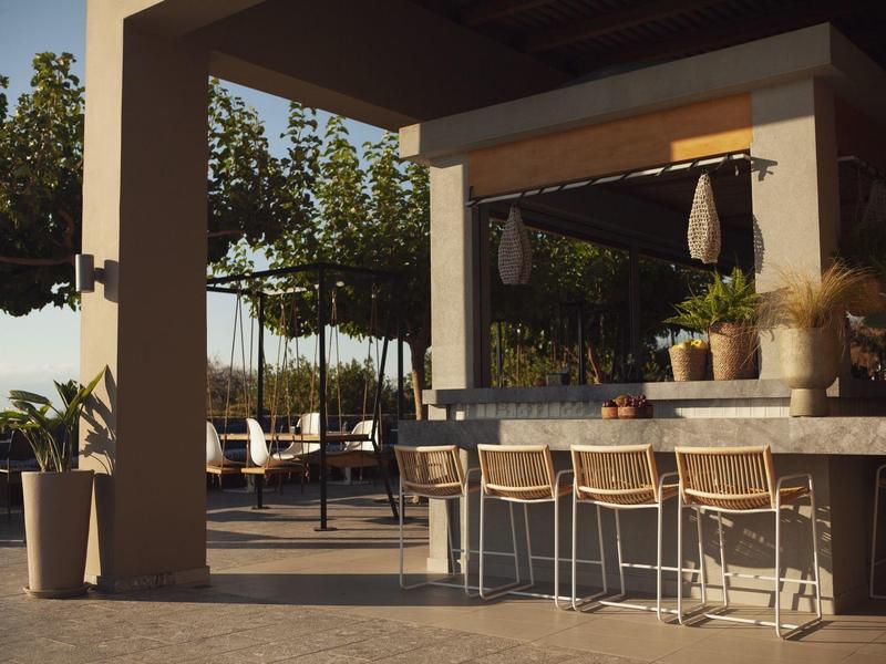 Zewnętrzny bar z krzesłami, roślinami i drzewami pod zadaszeniem o zachodzie słońca.