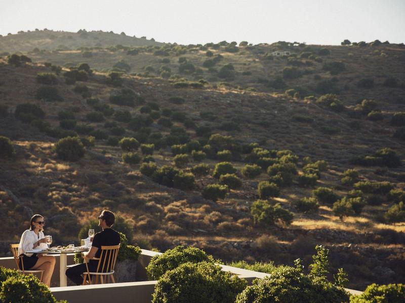 Dwie osoby jedzące na zewnątrz z widokiem na nasłonecznione wzgórze pokryte krzewami.