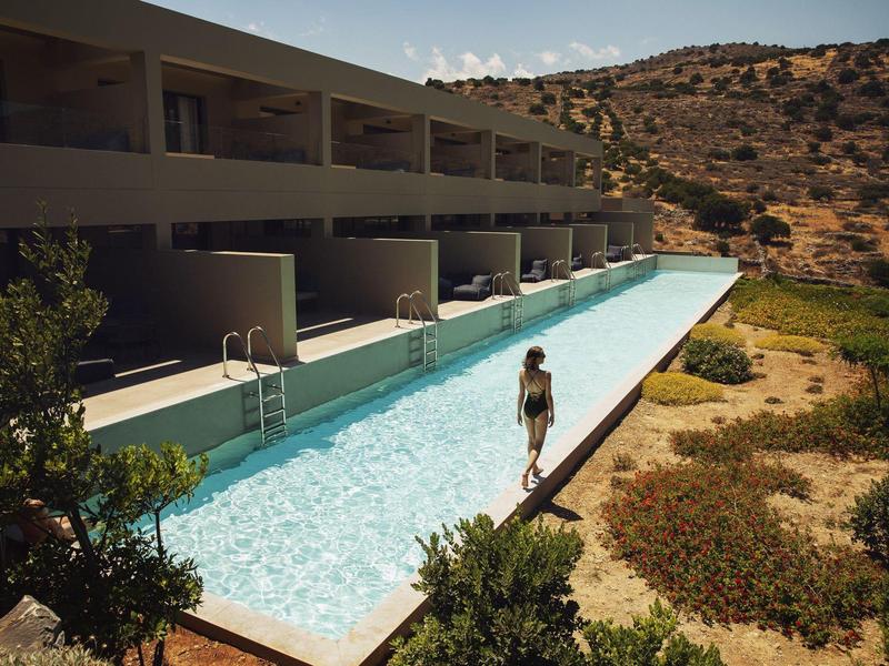 Długi basen na świeżym powietrzu obok nowoczesnego hotelu z widokiem na suche wzgórza i zieleń, z osobą spacerującą obok.