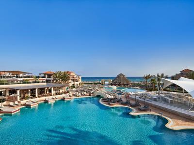 Velký hotelový bazén s venkovními terasami a výhledem na moře pod jasně modrou oblohou.