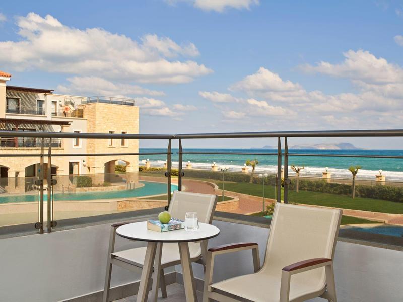 Balkon met stoelen en tafel met uitzicht op zee, zwembad en hotel onder een blauwe lucht.