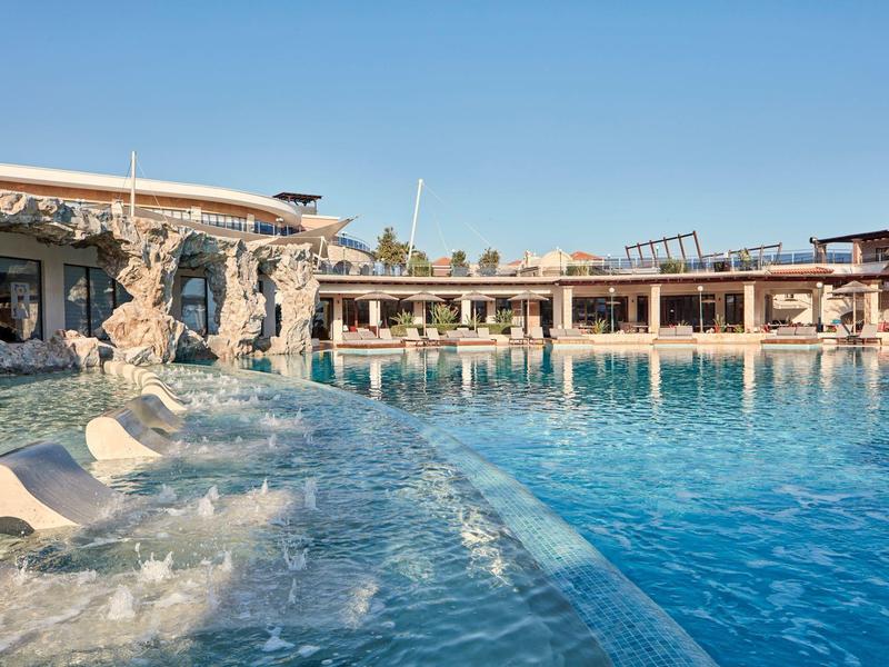 Modern hotel met groot zwembad en waterfonteinen onder een heldere lucht.