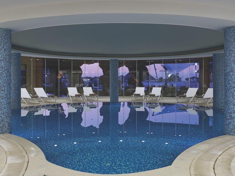 Overdekt zwembad met blauwe tegels, ligstoelen en grote glasramen