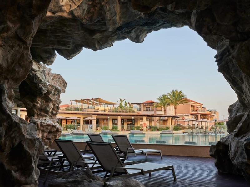 Uitzicht vanuit een rotsachtige grot op een zwembad en hotel met ligstoelen op de voorgrond.