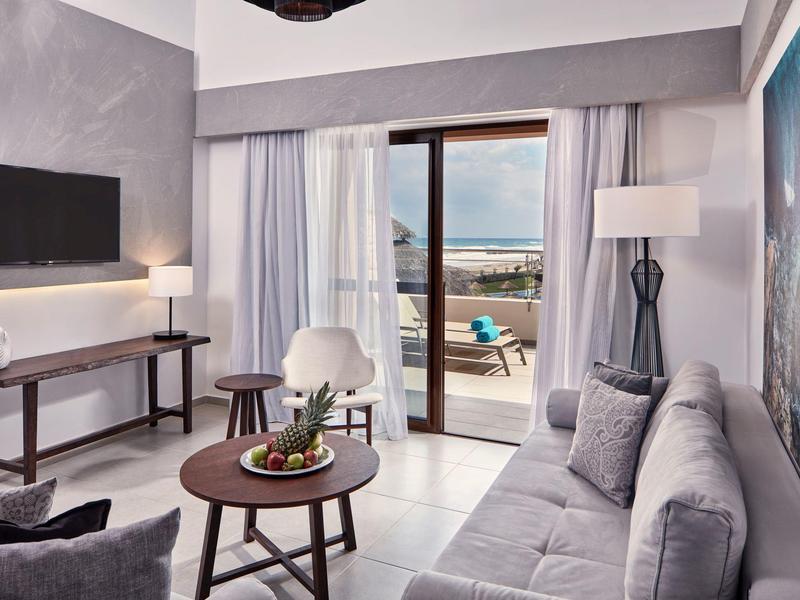 Modern ingerichte hotelkamer met bank, tv en terras met uitzicht op zee.