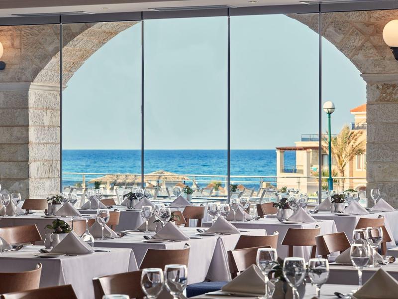 Restaurant met gedekte tafels bij grote ramen met uitzicht op zee en strand.