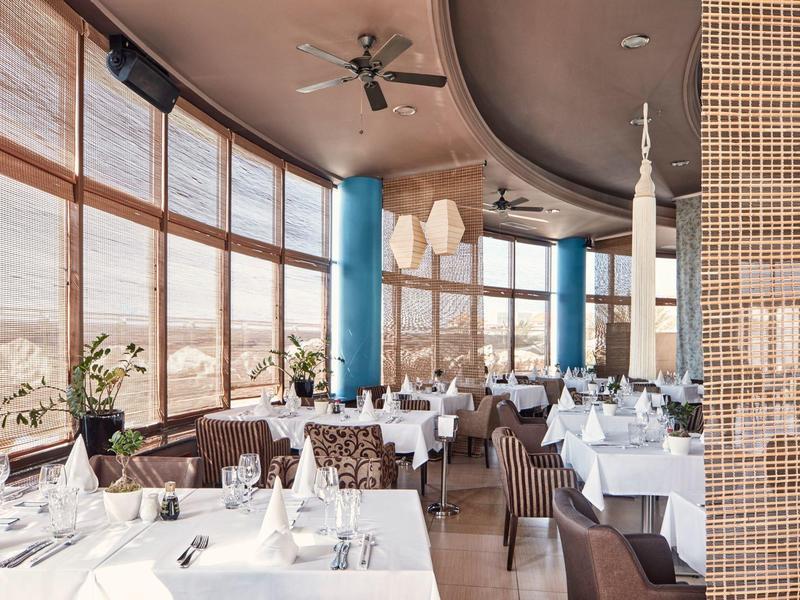 Elegant restaurant met grote ramen met uitzicht op zandduinen, gedekte tafels en plafondventilatoren.