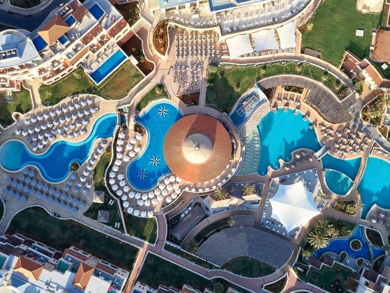 Luchtfoto van een hotel met meerdere gebogen blauwe zwembaden en parasols.