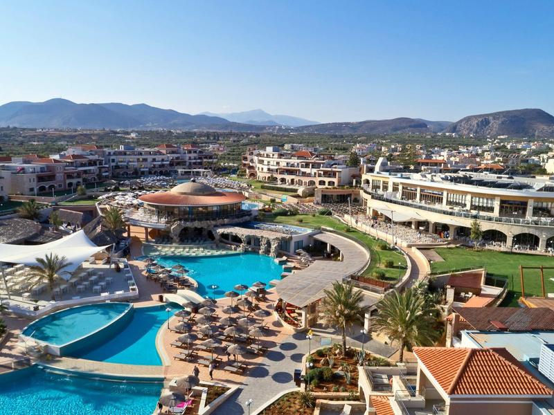Uitzicht op een resort met meerdere zwembaden, ligstoelen en omliggende gebouwen bij bergen.