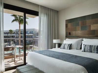 Chambre d'hôtel moderne avec balcon, grand lit et vue sur les bâtiments voisins et les palmiers.